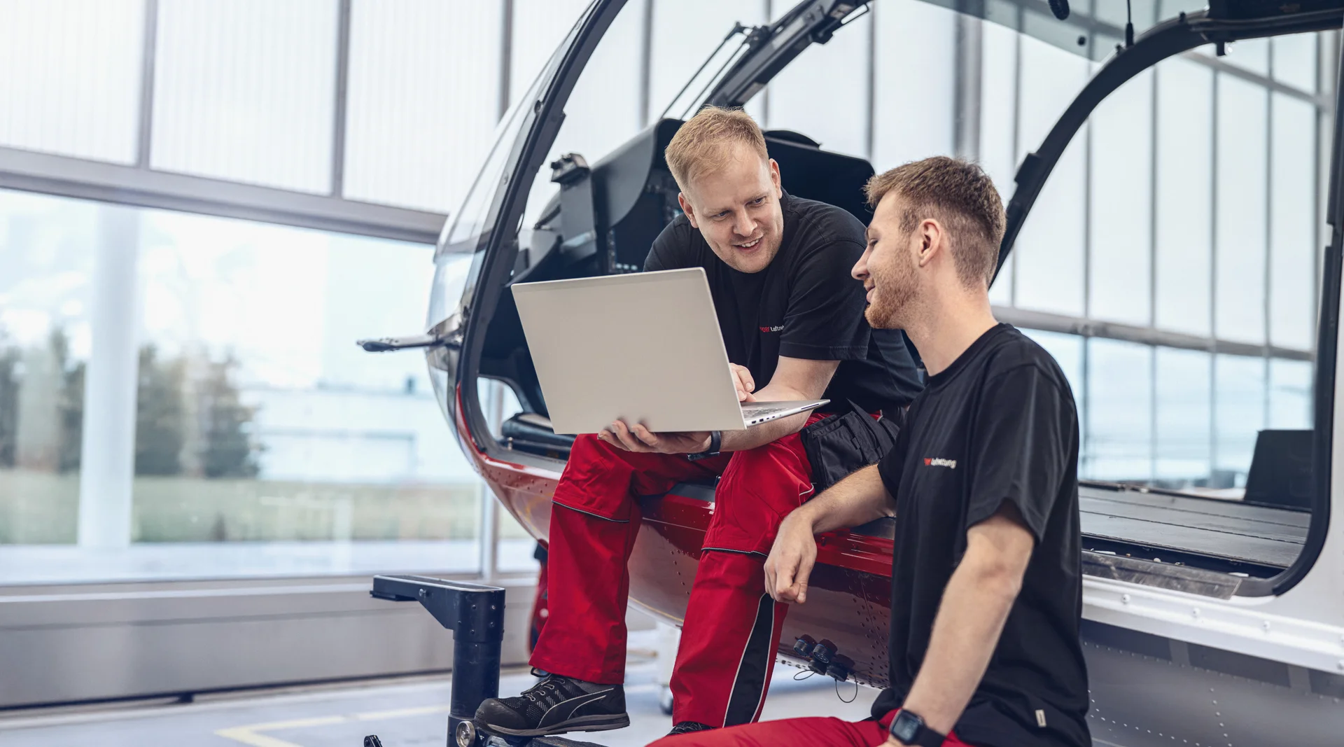 Zwei Techniker der DRF Luftrettung blicken auf einen Laptopbildschirm und sitzen dabei auf der Kufe eines Hubschraubers.